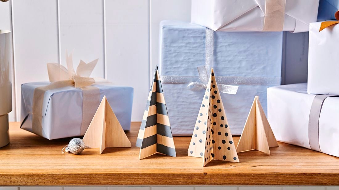 Four completed balsa wood Christmas tree figurines on a wooden table surrounded by Christmas presents