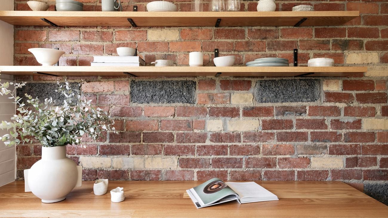 Brick wall in kitchen with wooden floating shelves and ceramic bowls sitting on top.