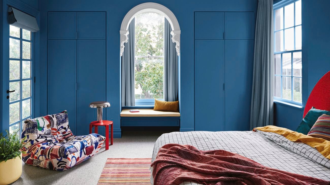 Bedroom painted depp blue with a colourful chair in left-hand corner and bed in right-hand corner