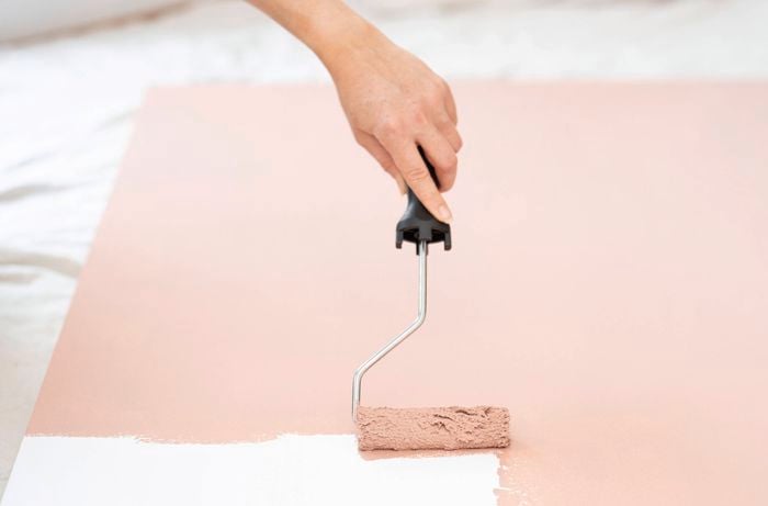 Applying pale pink paint to a wall panel with a paint roller as the first step in a DIY bookshelf project.
