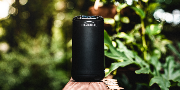 Thermacell portable mosquito repeller positioned on a timber railing in a lush outdoor garden setting.