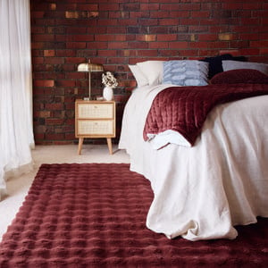A bedroom with a brick wall, bed, side table with a lamp on it and a burgundy rug on the floor in it.