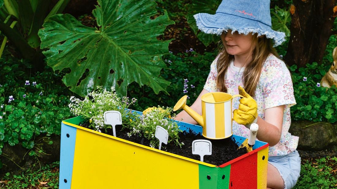 A child wearing a blue sunhat and gardening gloves kneels in a garden, planting seedlings in a brightly coloured raised planter box and watering them with a small yellow watering can