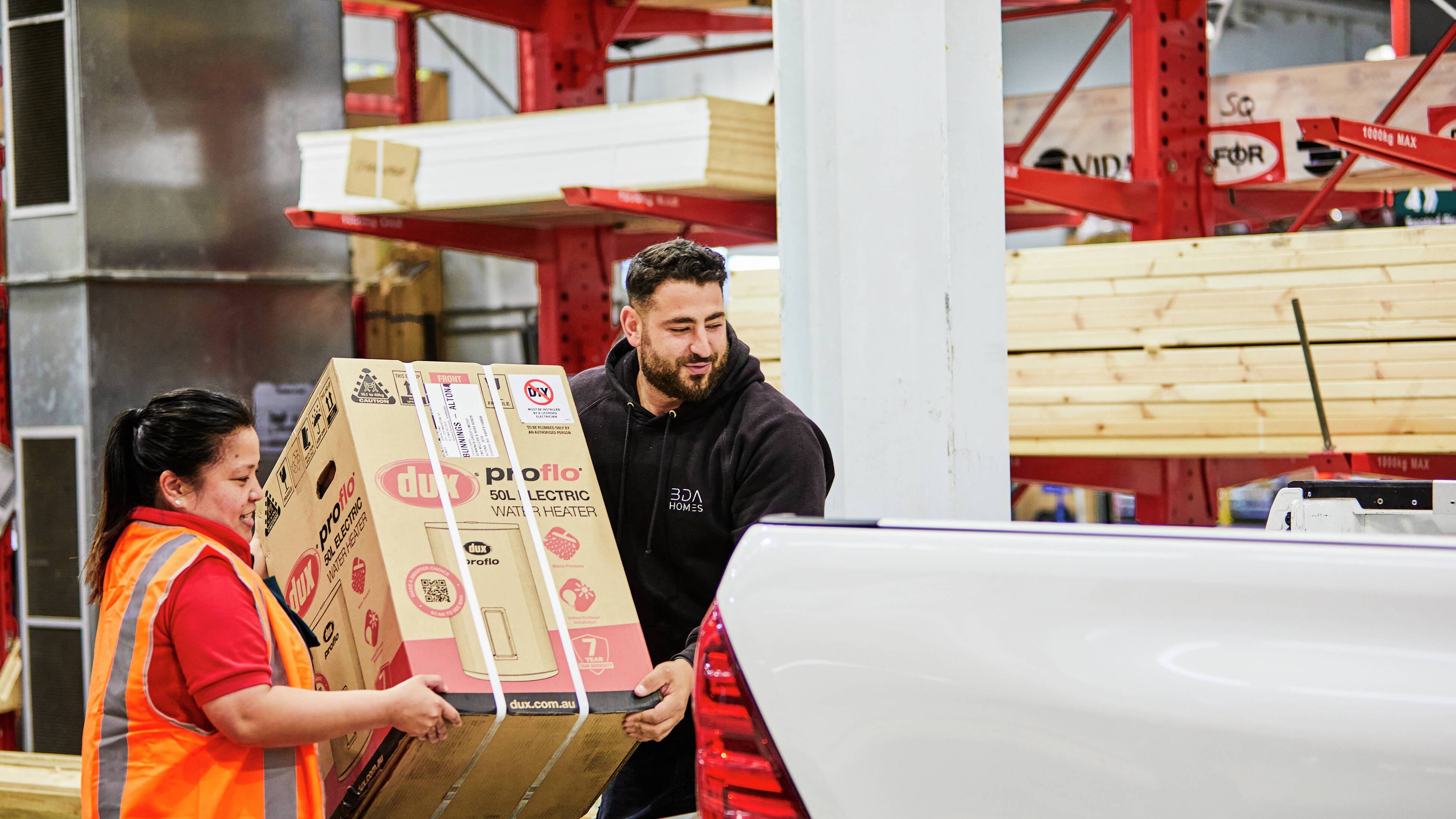 A female Bunnings Warehouse team member and a tradie load a hot water system into the back of a ute with timber in the background