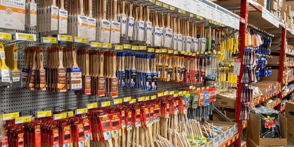 Bunnings aisle displaying a wide selection of paint brushes and decorating tools hanging on pegboard walls. Shelves are neatly stocked with brushes of various sizes, with yellow price labels running along the racks and boxed inventory stored on upper shelves.