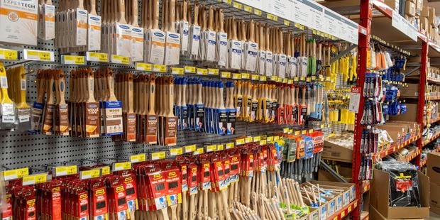 Bunnings aisle displaying a wide selection of paint brushes and decorating tools hanging on pegboard walls. Shelves are neatly stocked with brushes of various sizes, with yellow price labels running along the racks and boxed inventory stored on upper shelves.