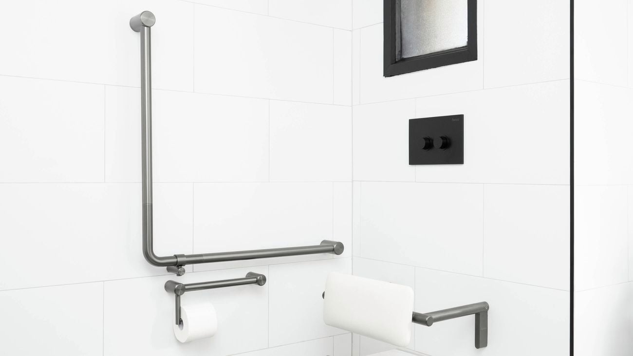 Close-up of a white tiled bathroom wall with stainless-steel L-shaped grab rail, horizontal grab rail, and a short rail with a toilet paper holder. A padded white backrest is mounted beside them.