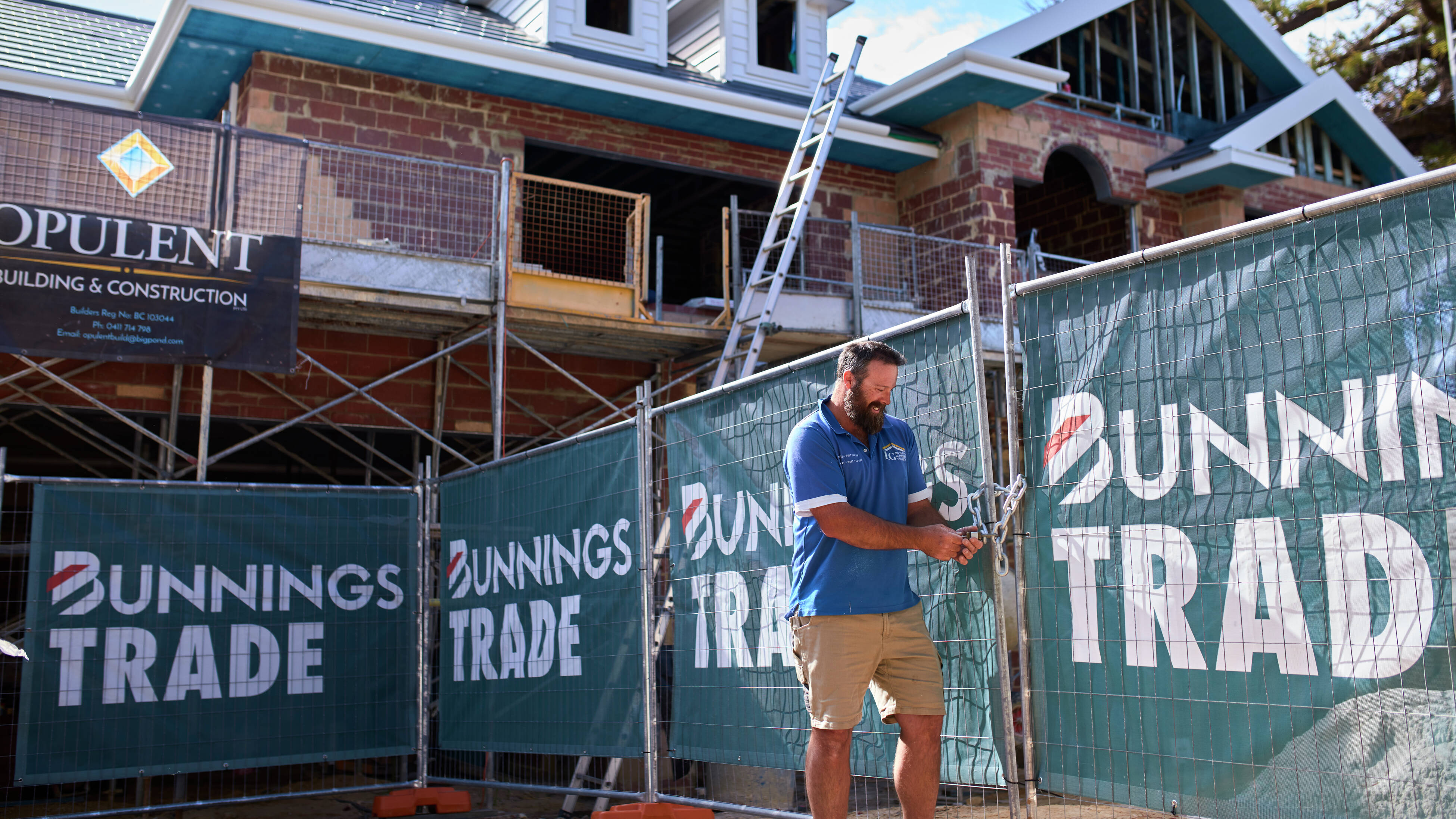 A male tradie wearing a blue shirt stands next to a construction site fence wrapped in Bunnings Trade mesh while locking up the site