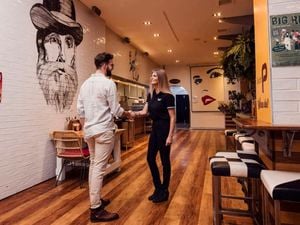 Two people shaking hands inside a modern café or restaurant with wooden floors, wall murals, bar seating, and posters visible along the walls.