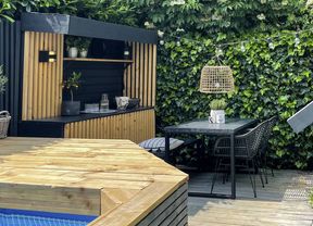 A timber deck with a table and chairs, built-in buffet and the edge of a timber-framed pool in the foreground and a shed to the right.