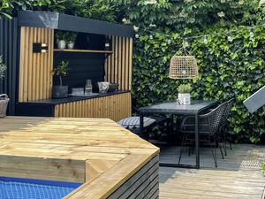A timber deck with a table and chairs, built-in buffet and the edge of a timber-framed pool in the foreground and a shed to the right.
