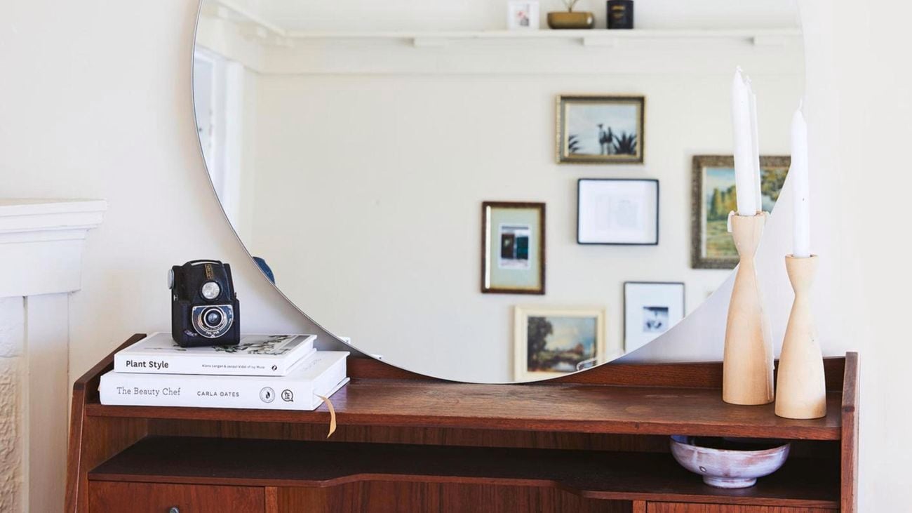 A shelf with a mirror, camera, books and candles on top. In the reflection of the mirror are hanging picture frames.