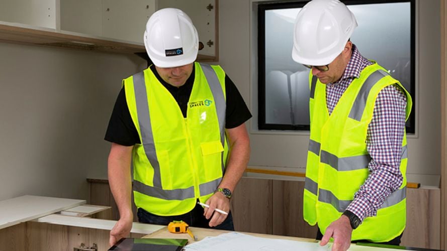 two work man in green high vis tops hearing white hard hats reviewing kitchen