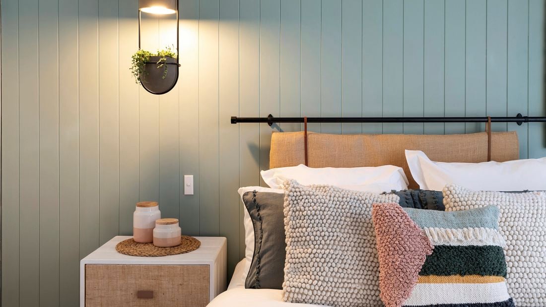 Bed with DIY hessian bedhead and lots of decorative pillows against a green panelled wall.