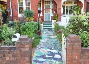 Front garden of a red‑brick period home with a white gate and a metal arch over a front path with a painted mural. Dense plants line the path, leading to a front porch with green tiled steps and a white security door.