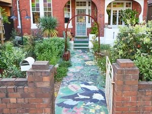 Front garden of a red‑brick period home with a white gate and a metal arch over a front path with a painted mural. Dense plants line the path, leading to a front porch with green tiled steps and a white security door.