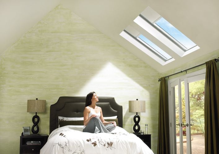 Calm-looking woman sitting on a bed with her eyes closed, facing an angled ceiling with two skylights with roller blinds