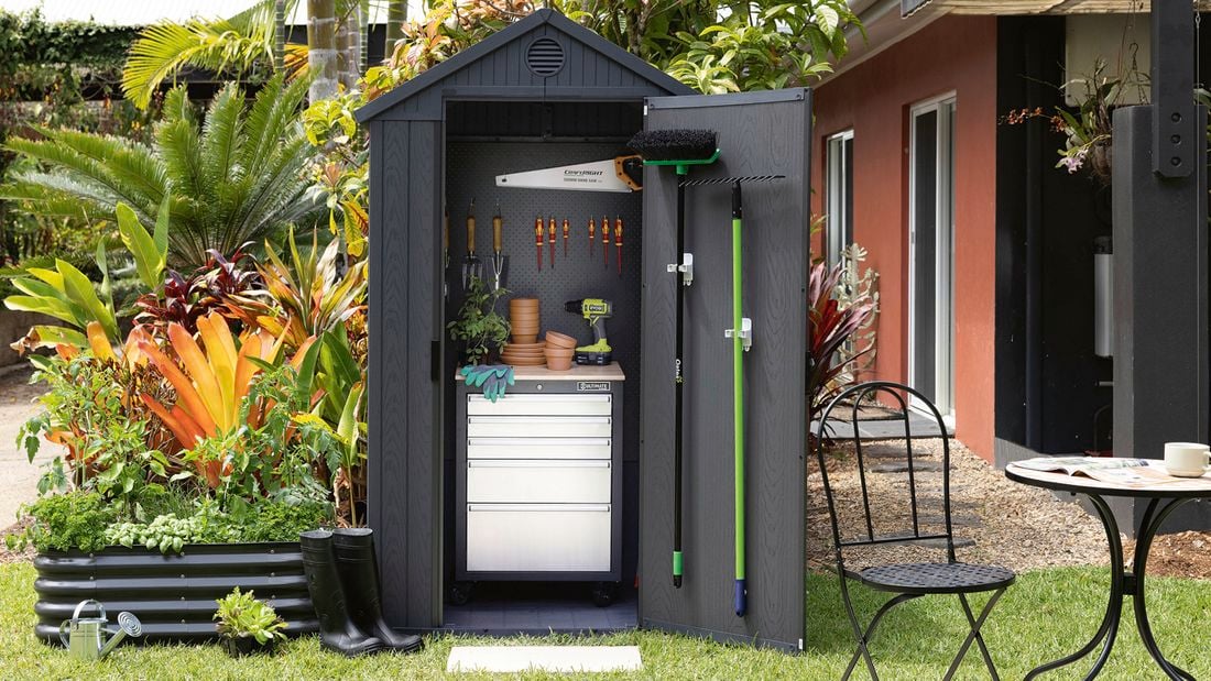 A small dark grey shed with its door open to show a drawer cabinet, tools on a pegboard and a rake and broom hanging on the door