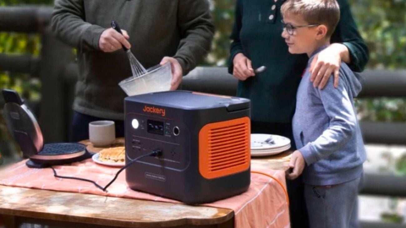 A family stands around a table outdoors with a Jackery portable power station, waffle maker, and food items.