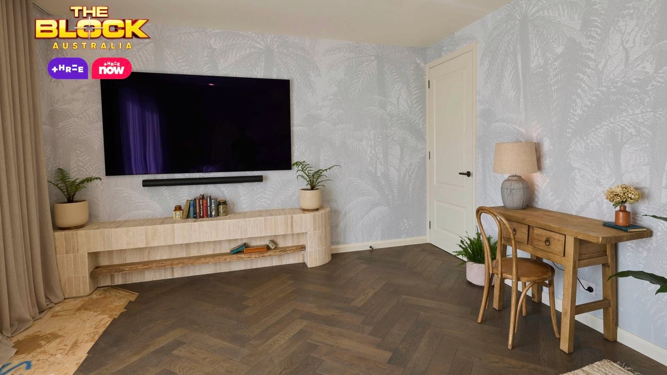 A modern living room featuring a wall-mounted flat-screen TV above a wooden shelf with books and decorative items. To the right, a wooden desk with a chair, lamp, and plants sits against light-patterned wallpaper. The floor has a herringbone design.