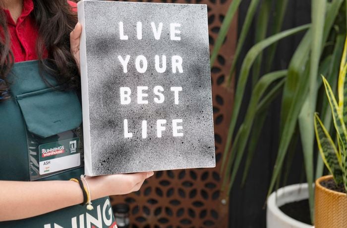 Person holding up a canvas with the stencilled words "Live your best life"