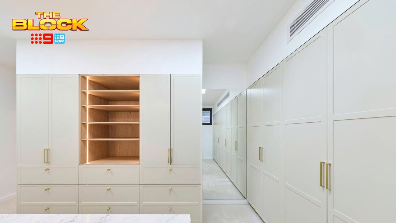Bright white walk-in wardrobe with built-in cabinetry, timber shelving, gold handles, drawers and a large mirrored panel.