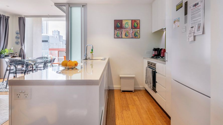 Modern white kitchen with a stone island bench, built-in appliances, timber flooring and an open-plan dining area leading to a balcony with city views.
