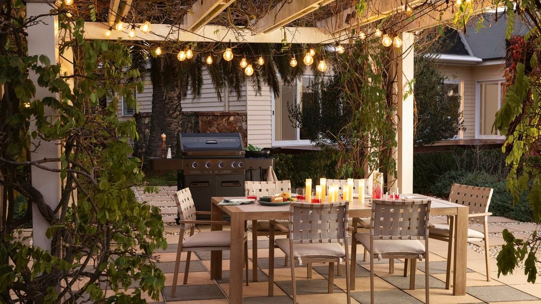   Outdoor dining area under a pergola decorated with hanging string lights, featuring a wooden table set with candles, plates, and drinks, surrounded by woven-back chairs on a tiled patio, with a barbecue grill in the background and greenery framing the space.