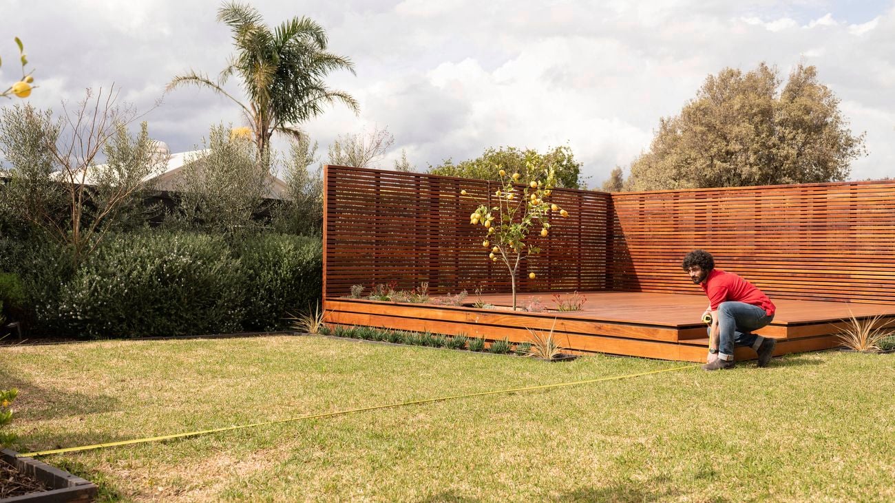 A Bunnings Team Member is measuring the length of a lawn using a tape measure near a wooden deck in a garden.
