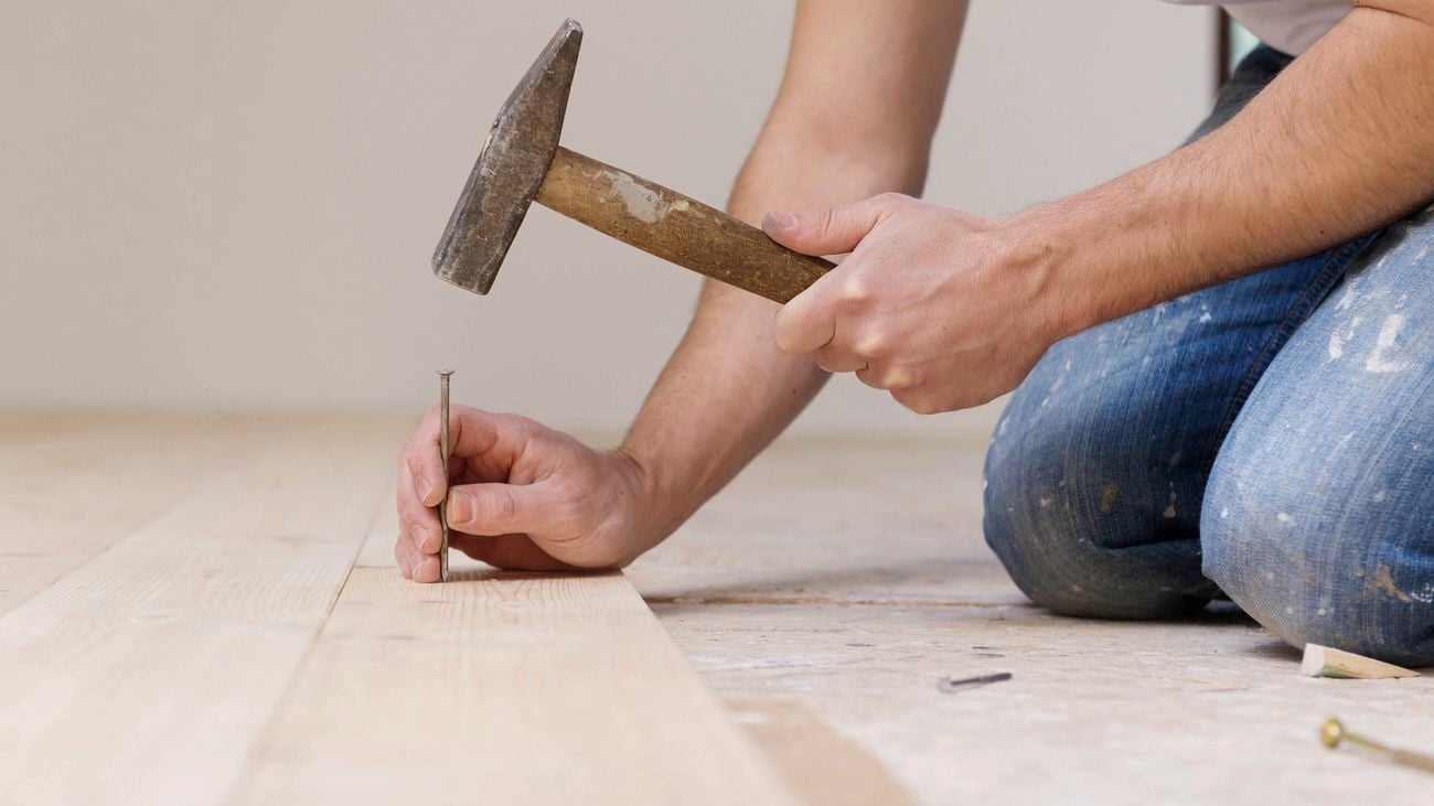 Hands hammering a nail into a wooden floorboard.