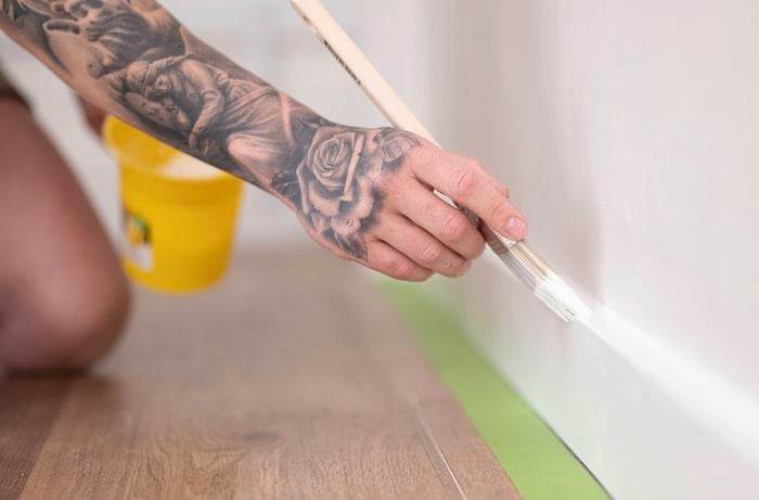 A person is painting a skirting board white with a small paint brush, while using painter's tape to protect the floor.