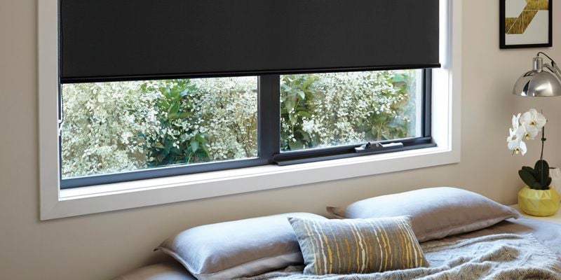 A black roller blind installed in a window in a light themed bedroom
