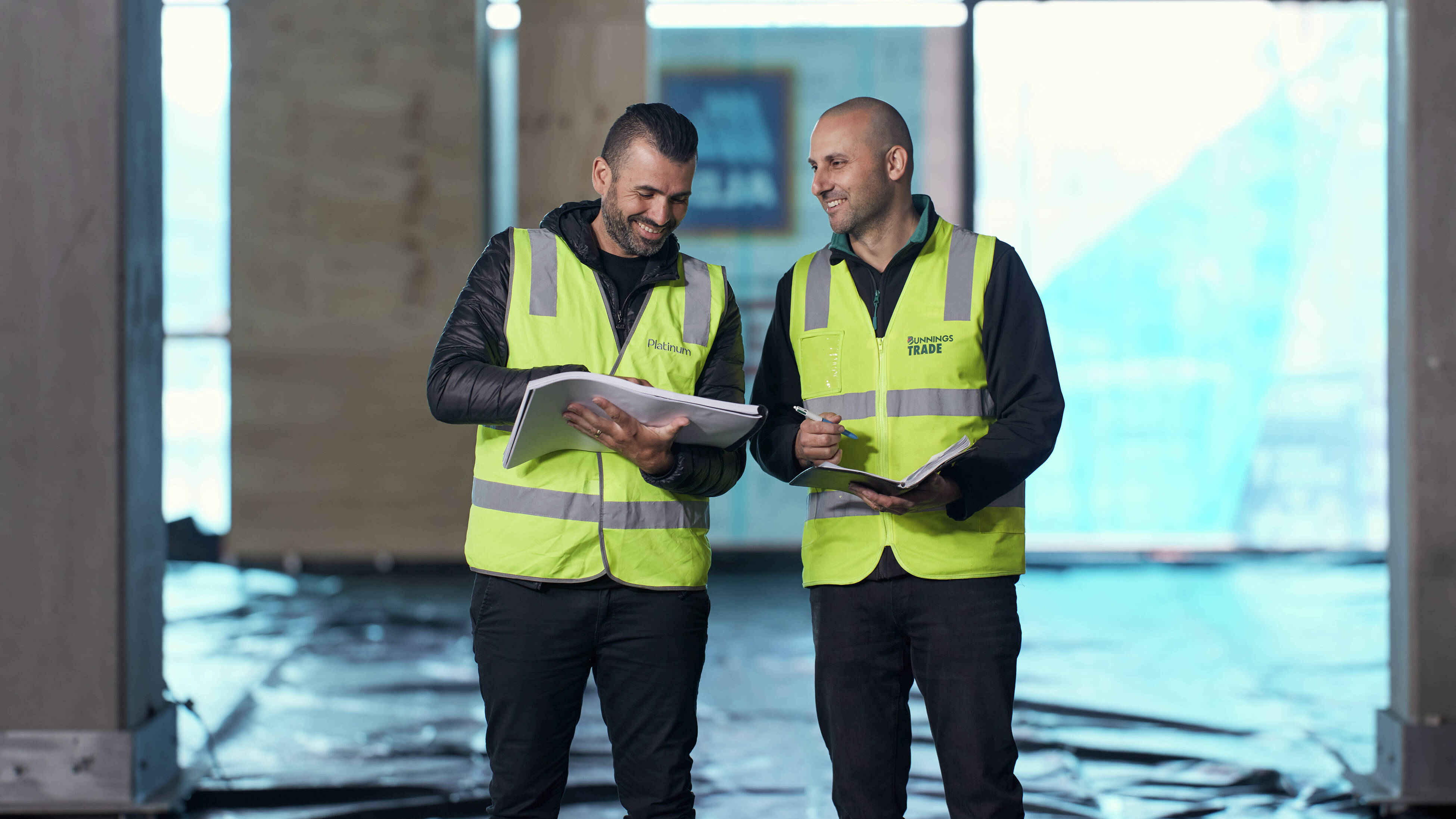 Two men standing on an indoor construction site, one holding plans and the other a checklist in a notebook.