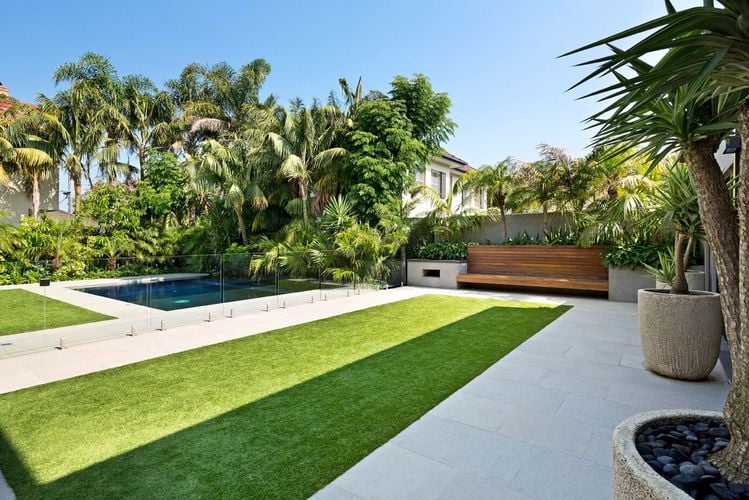 A narrow strip of lawn in front of a wooden and stone bench overlooking a pool area