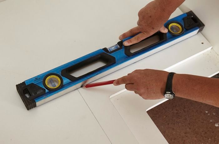 A person using a spirit level as a straight edge to draw a line on a cabinet panel