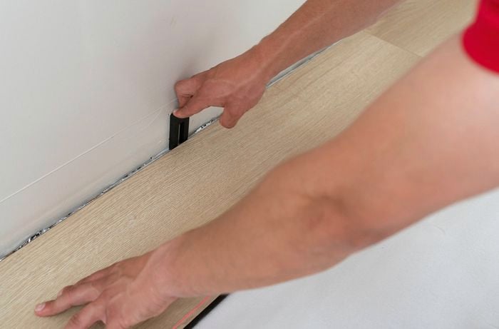A person is laying the first laminate floorboard with a packer to ensure proper wall alignment.