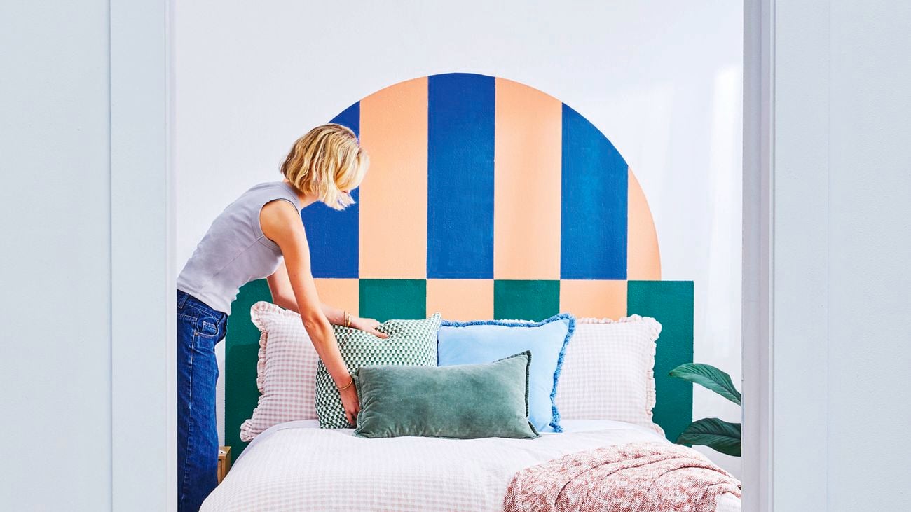 A person with blonde hair in a white top and jeans arranges colourful pillows on a bed with a geometric blue, apricot, and green D.I.Y. bed headboard
