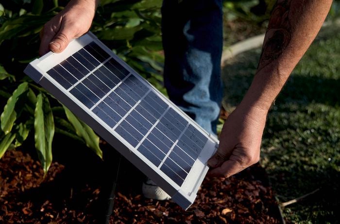 Hands hold a solar panel above a garden bed with green plants.
