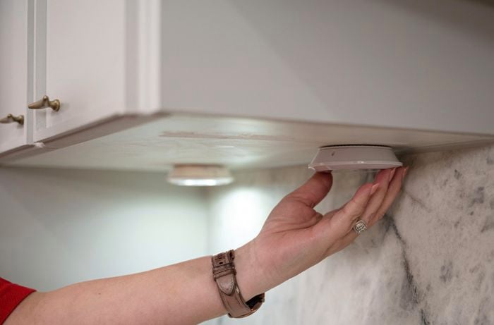 A person's hand reaching out to touch undermount lights installed beneath a kitchen cabinet.