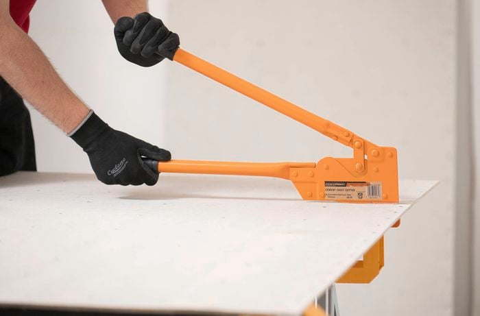 A person wearing gloves is using an orange cement sheet cutter to cut an underlay sheet to size.