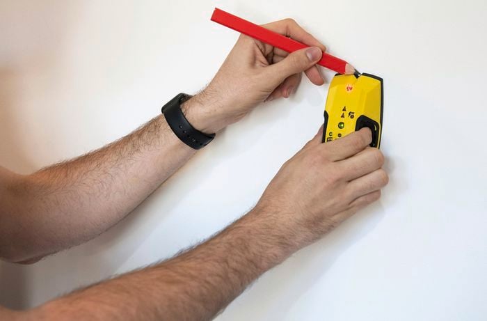 A stud finder being used to locate the wall stud on a white wall, and a pencil being used to mark its position