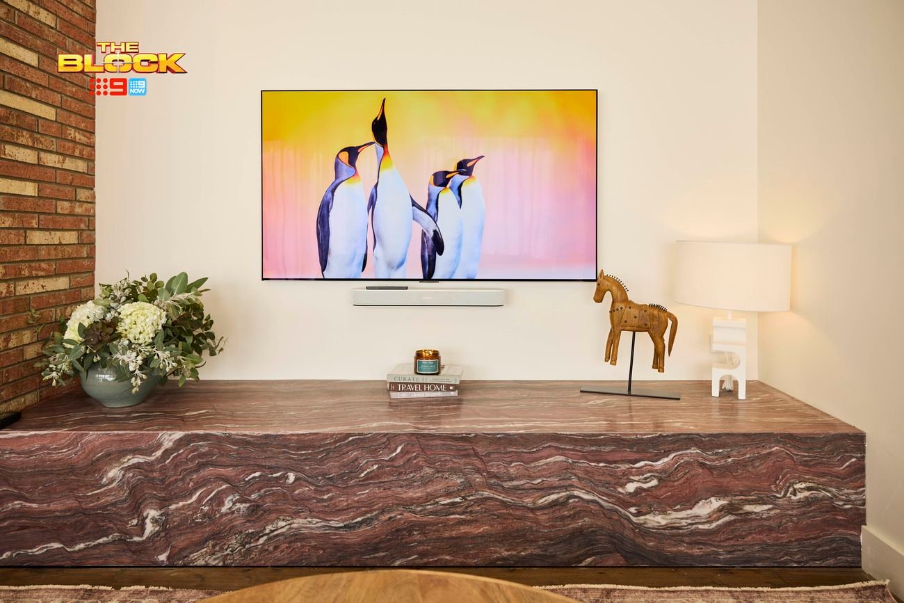 A modern living room featuring a central brick fireplace, exposed wooden ceiling beams, and a large abstract painting on the right wall. Furnished with a brown leather sofa, two gray armchairs, and a round wooden coffee table atop an area rug. A dining table set for a meal is visible to the left. 