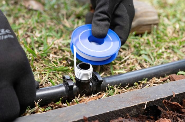 A person is connecting black pipes using a barbed and threaded tees on grass next to a concrete slab.