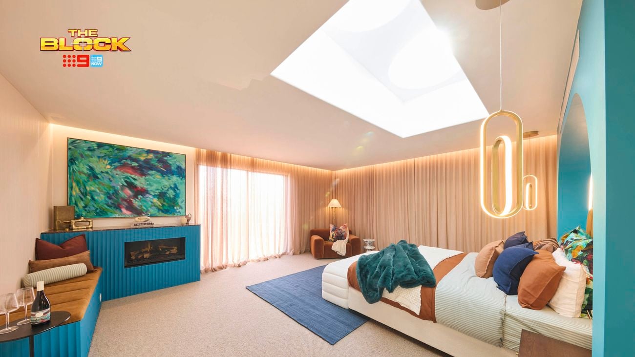 A modern bedroom with a large skylight, featuring a bed with multiple pillows and blankets, a blue rug, and a side table with wine glasses. An abstract painting hangs above a fireplace on the left wall. The room has beige carpeting, curtains on the far wall, a unique hanging light fixture near the bed, and a seating area in the corner. 
