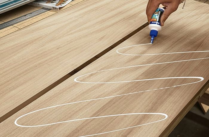 Glue is applied in a zigzag pattern to the surface of a timber panel on a workbench.