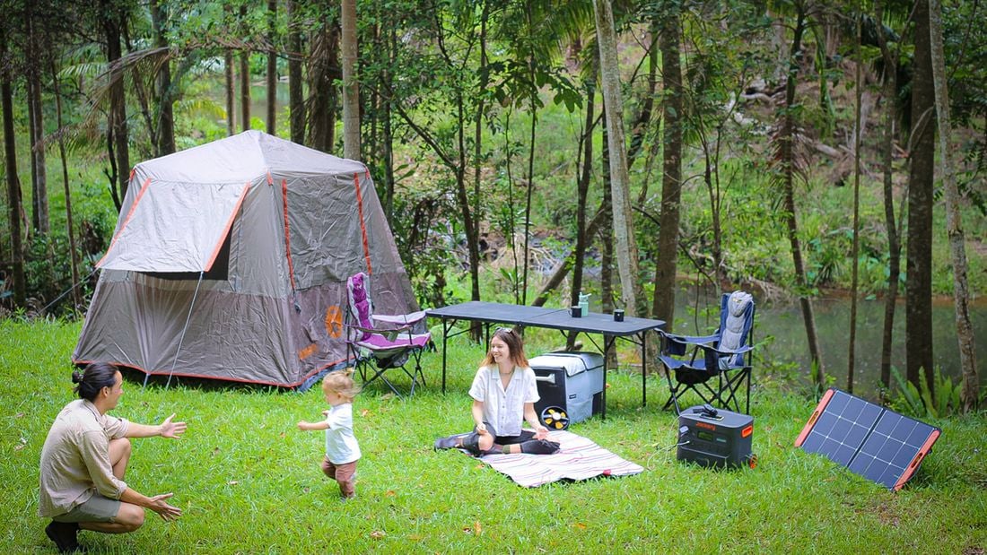 A camping setup in the bush with a tent, chairs, a table, a Jackery power station and a solar panel. A man kneels reaching towards a toddler, while a woman sits on a picnic mat.