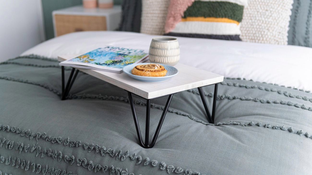 White painted bed tray topped with a magazine, danish on a plate and mug on a made up bed