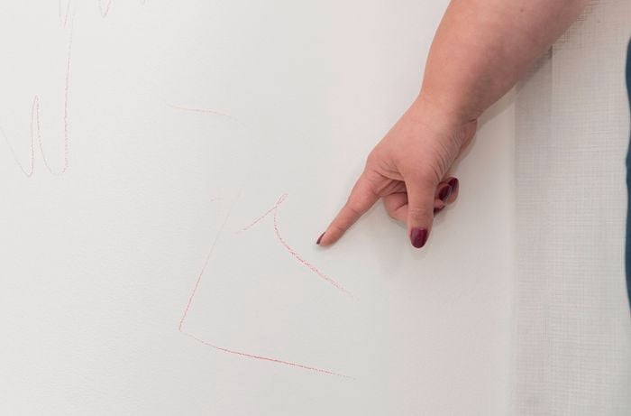A hand with painted nails points to red crayon marks drawn on a white wall.