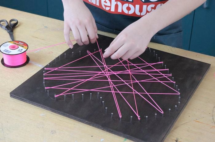 Person threading pink string around nails in plywood sheet