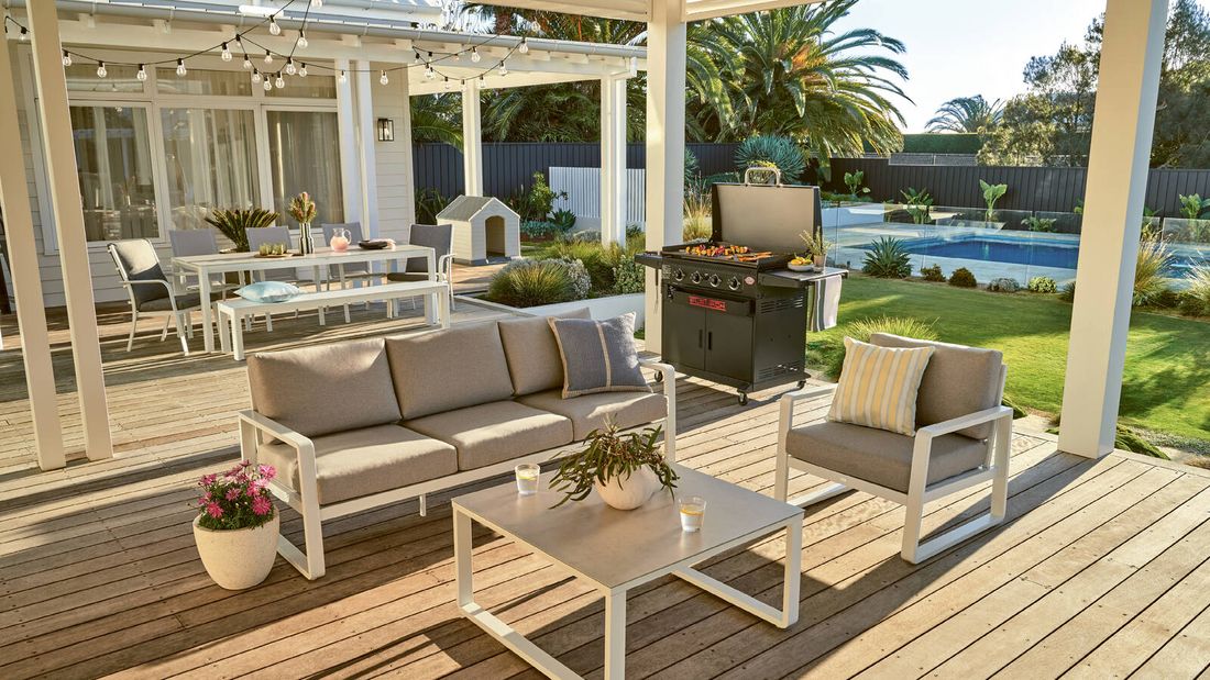 Contemporary deck with outdoor lounge furniture, a dining setup under string lights, a barbie cooking food, and a garden with a pool.